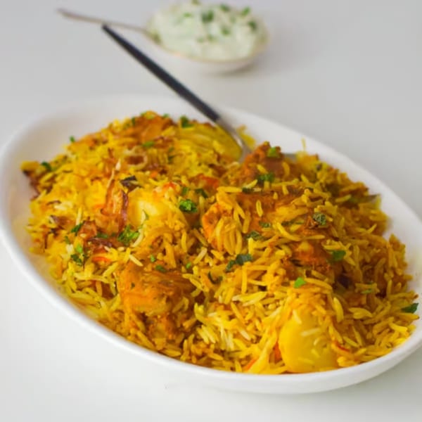 A white oval dish filled with vibrant yellow chicken biryani, garnished with fresh green herbs.
