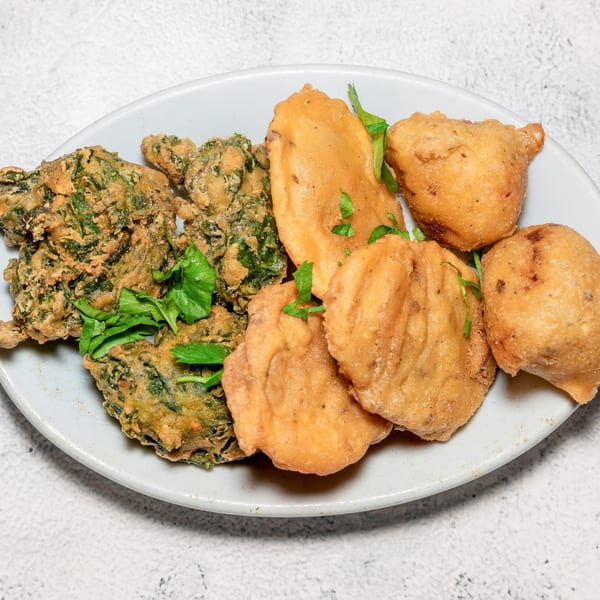 A white plate holds various Indian pakoras, some green and leafy, others golden brown, garnished with fresh herbs.