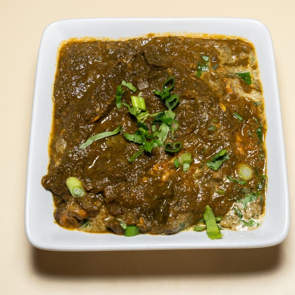 A white square bowl filled with a dark green, creamy Indian dish, garnished with fresh herbs.